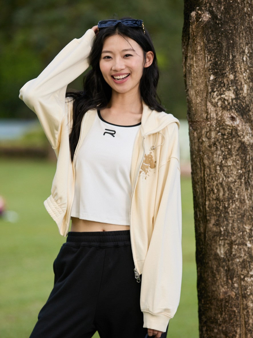 Smiling Asian female model standing by a tree with one hand over her head, wearing a khaki Ramsports EcoSoft Logo Hoodie, black pants, and a white top