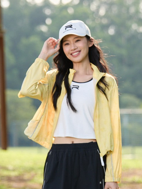 Smiling Asian female model wearing a yellow Ramsports FlexTek Marble Print Jacket over a white top and black pants, holding the white cap she’s wearing as she's looking at the camera