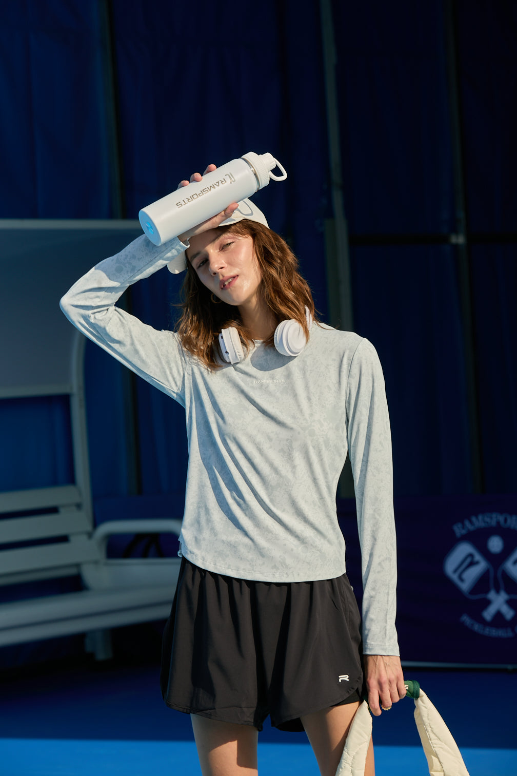 Female model wiping sweat while holding a tumbler, wearing gray long sleeves, FlexTek Pace Shorts in black, and headphones around her neck
