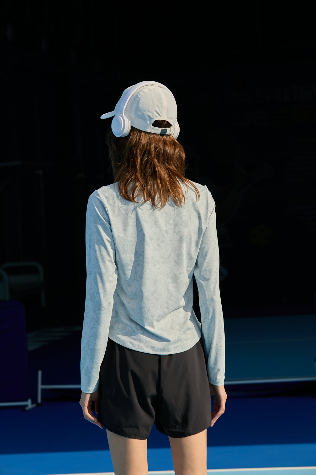 Back view of female model in white cap with headphones, gray long sleeves, and FlexTek Pace Shorts in black.