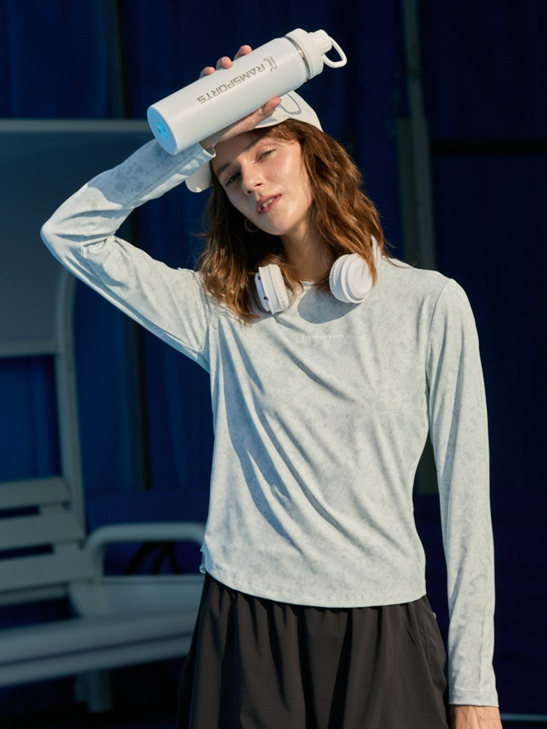 Female model wearing a gray Ramsports CoolDry Marble Print Long-Sleeves Tee and black shorts, posing with one hand on top of her cap as if wiping sweat, while holding a tumbler