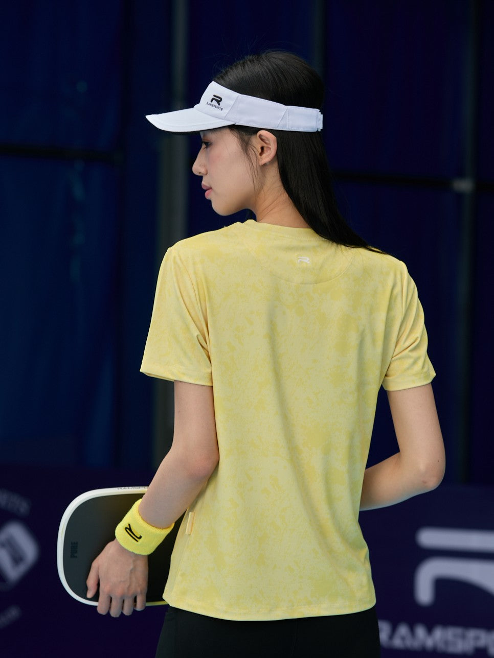 Asian female model facing away, wearing a yellow Ramsports CoolDry Marble Print Tee, black shorts, a yellow wristband, and a white visor while holding a paddle