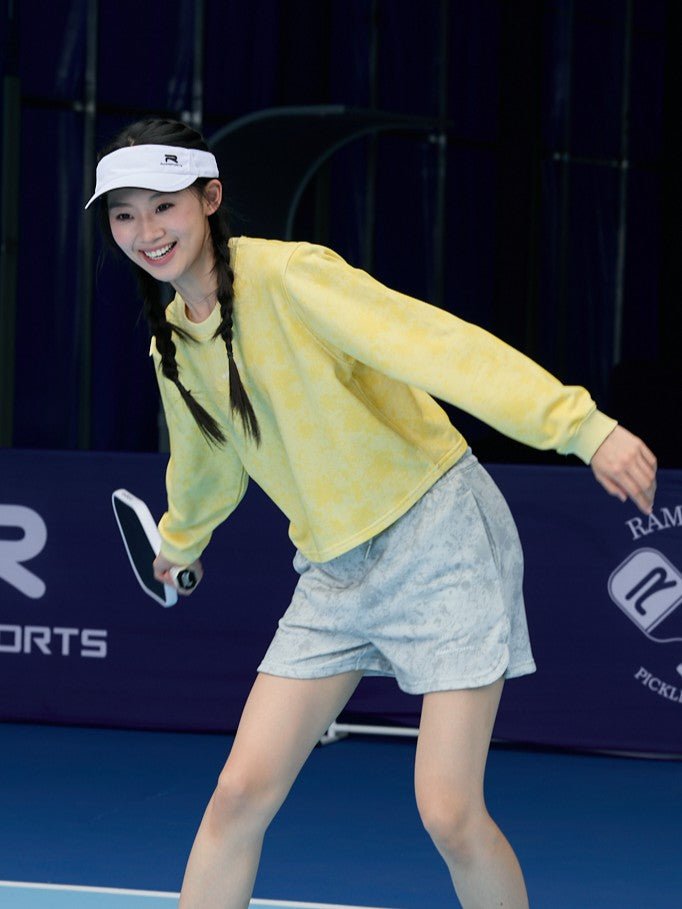 Smiling Asian female model in a yellow long-sleeve top and gray Ramsports EcoSoft Marble Print Shorts, posed as if playing pickleball with a paddle in hand
