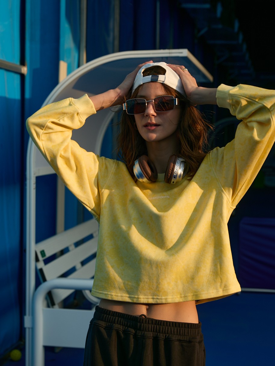 Female model holding her head over her cap, wearing a yellow Ramsports EcoSoft Marble Print Crop Sweatshirt and black bottoms
