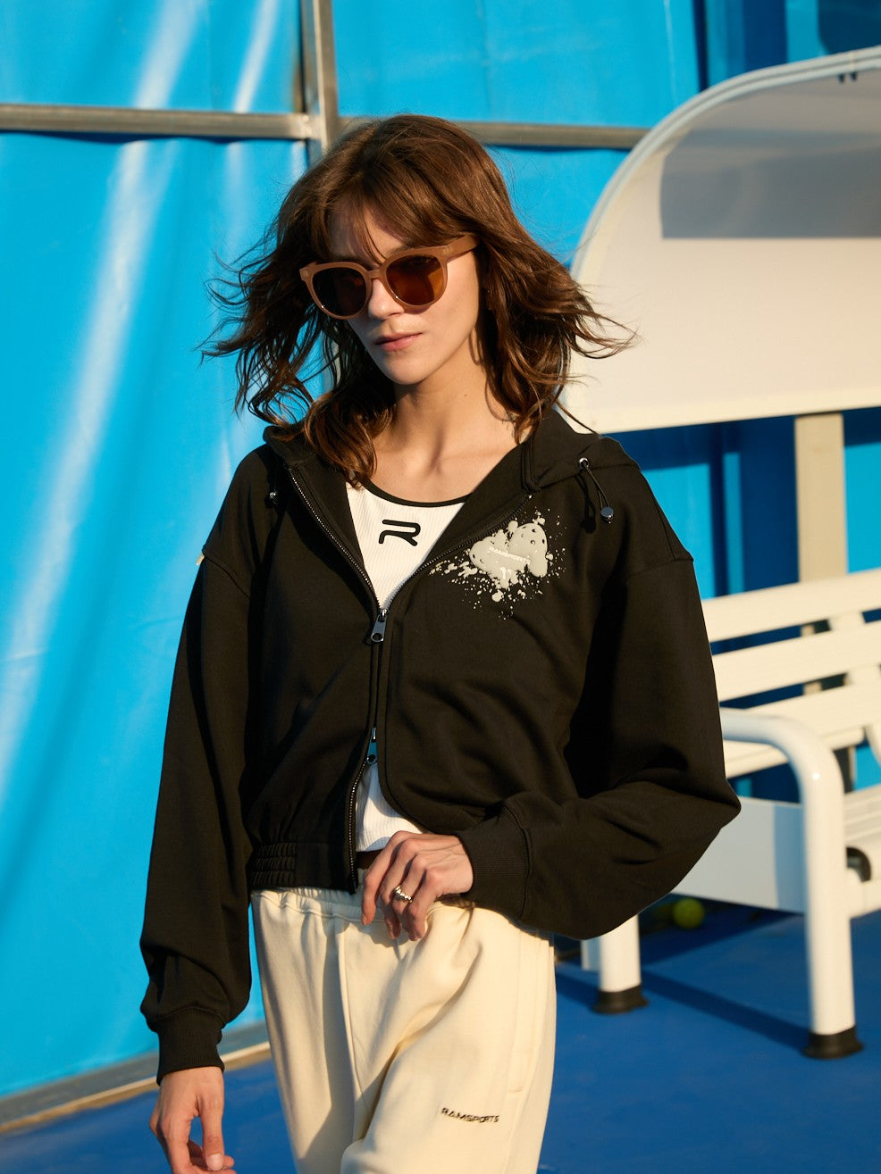 Female model wearing a black Ramsports EcoSoft Logo Hoodie over a white top, with the hood down and one hand resting on her pants