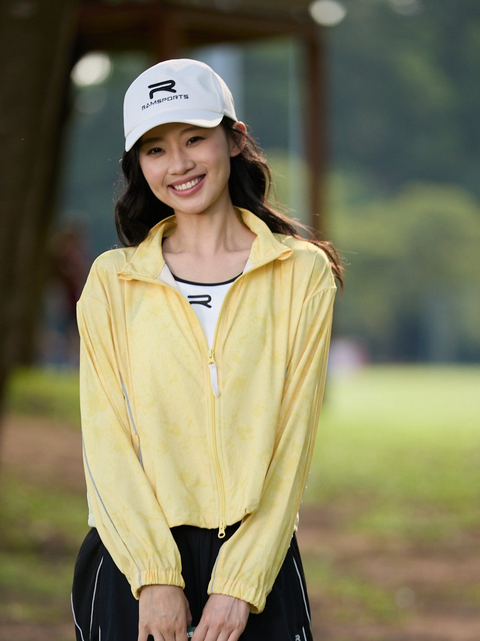 Smiling Asian female model wearing a yellow Ramsports FlexTek Marble Print Jacket over a white top and black pants
