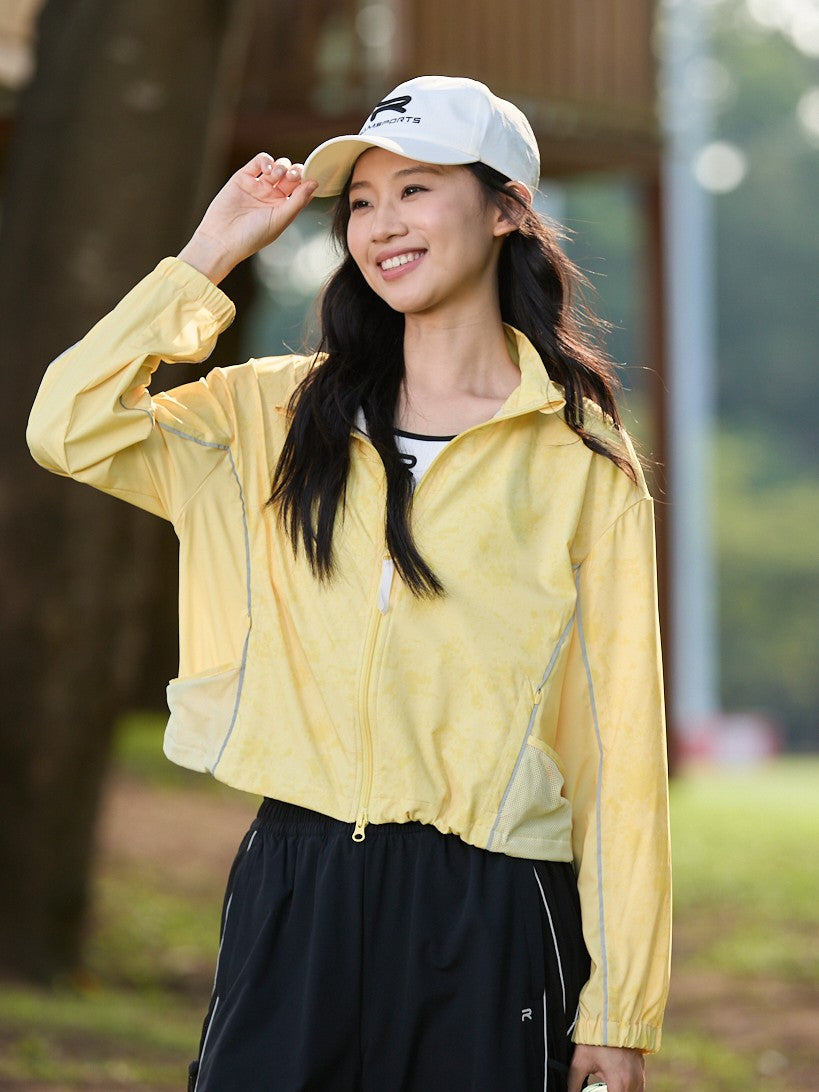 Smiling Asian female model wearing a yellow Ramsports FlexTek Marble Print Jacket over a white top and black pants, holding the white cap she’s wearing as she's looking away